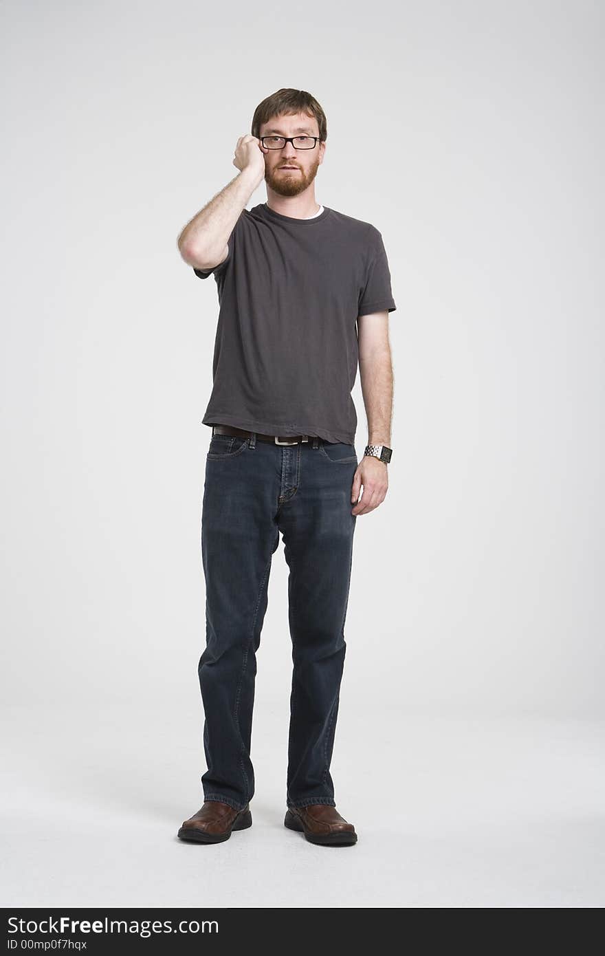 Man with beard wearing a gray T-shirt and denim pants stands.  He stands with straight with one hand brushing his cheek. Man with beard wearing a gray T-shirt and denim pants stands.  He stands with straight with one hand brushing his cheek.