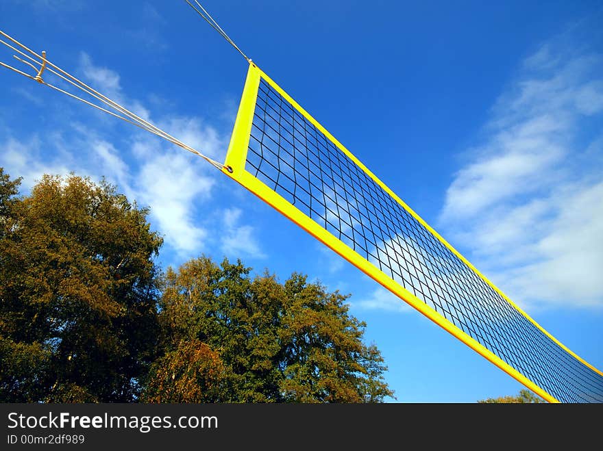 Yellow volleyball net under blue clouded sky, in the background autumn trees