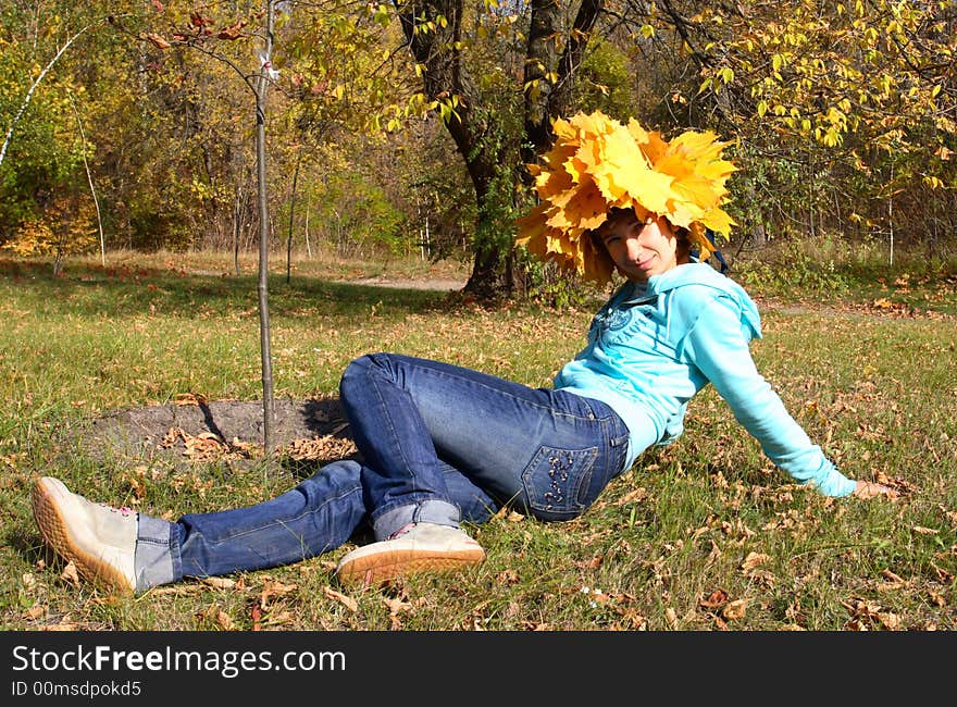 A girl in a chaplet from yellow maple leaves sits on a grass looks in a lens