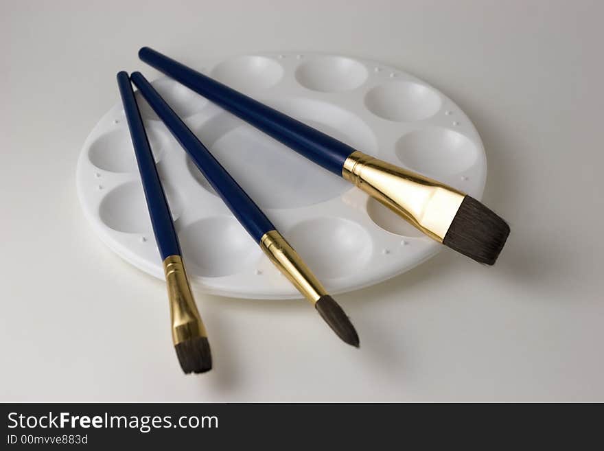 Three blue artist brushes on top of an empty paint palette with a white background. Three blue artist brushes on top of an empty paint palette with a white background.