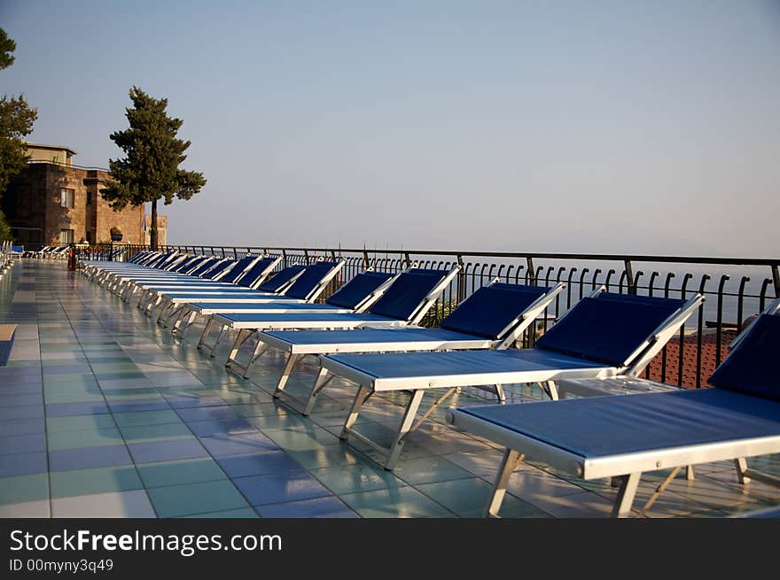 Empty Sun Loungers arranged in a neat row are highlighted by the early morning sunshine. Colored tiles around the swimming pool frame the photo. Empty Sun Loungers arranged in a neat row are highlighted by the early morning sunshine. Colored tiles around the swimming pool frame the photo