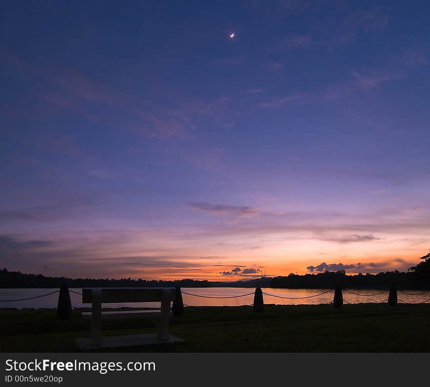 A bright crescent moon high in the twilight sky above an empty stone bench in a public park \. A bright crescent moon high in the twilight sky above an empty stone bench in a public park \