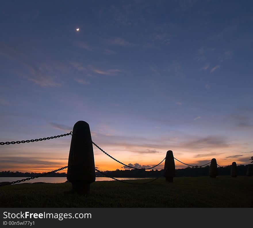 A bright crescent moon high in the twilight sky framed by a chain fence on the shores of a lake. A bright crescent moon high in the twilight sky framed by a chain fence on the shores of a lake