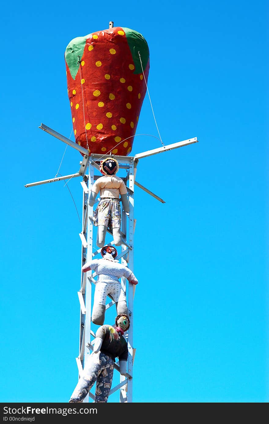 Three scarecrows hanging from a metal structure with a red strawberry on top of the structure. Three scarecrows hanging from a metal structure with a red strawberry on top of the structure