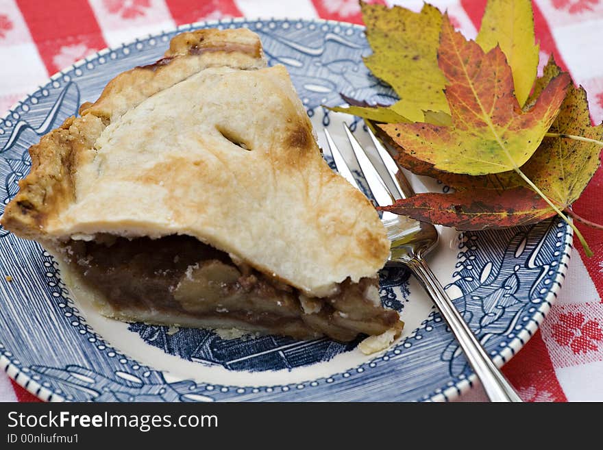 Slice of apple pie on old blue china and  gingham tablecloth.  Fall leaves on side. Slice of apple pie on old blue china and  gingham tablecloth.  Fall leaves on side.