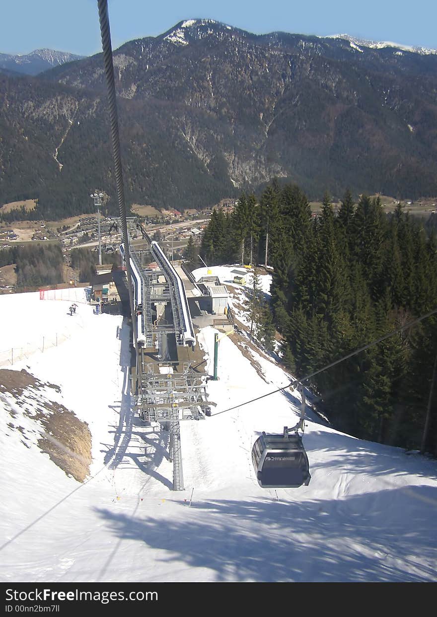 View from the cabin of the ski lift station and one funifor cabin going down, two skiers waiting, skiing resort in the valley, mountain background