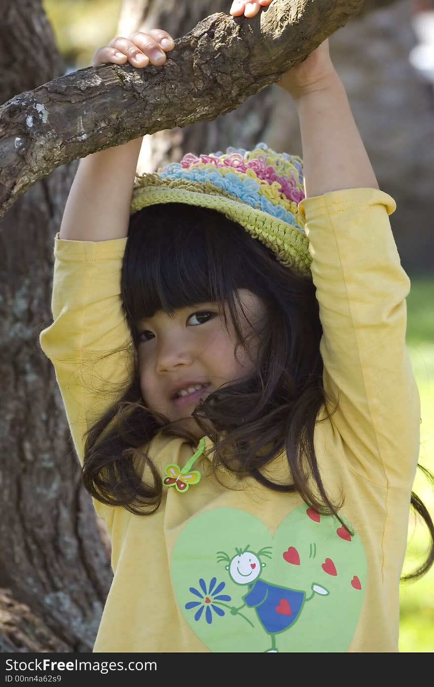 Cute little girl in yellow top and rainbow hat hanging from a tree. Cute little girl in yellow top and rainbow hat hanging from a tree