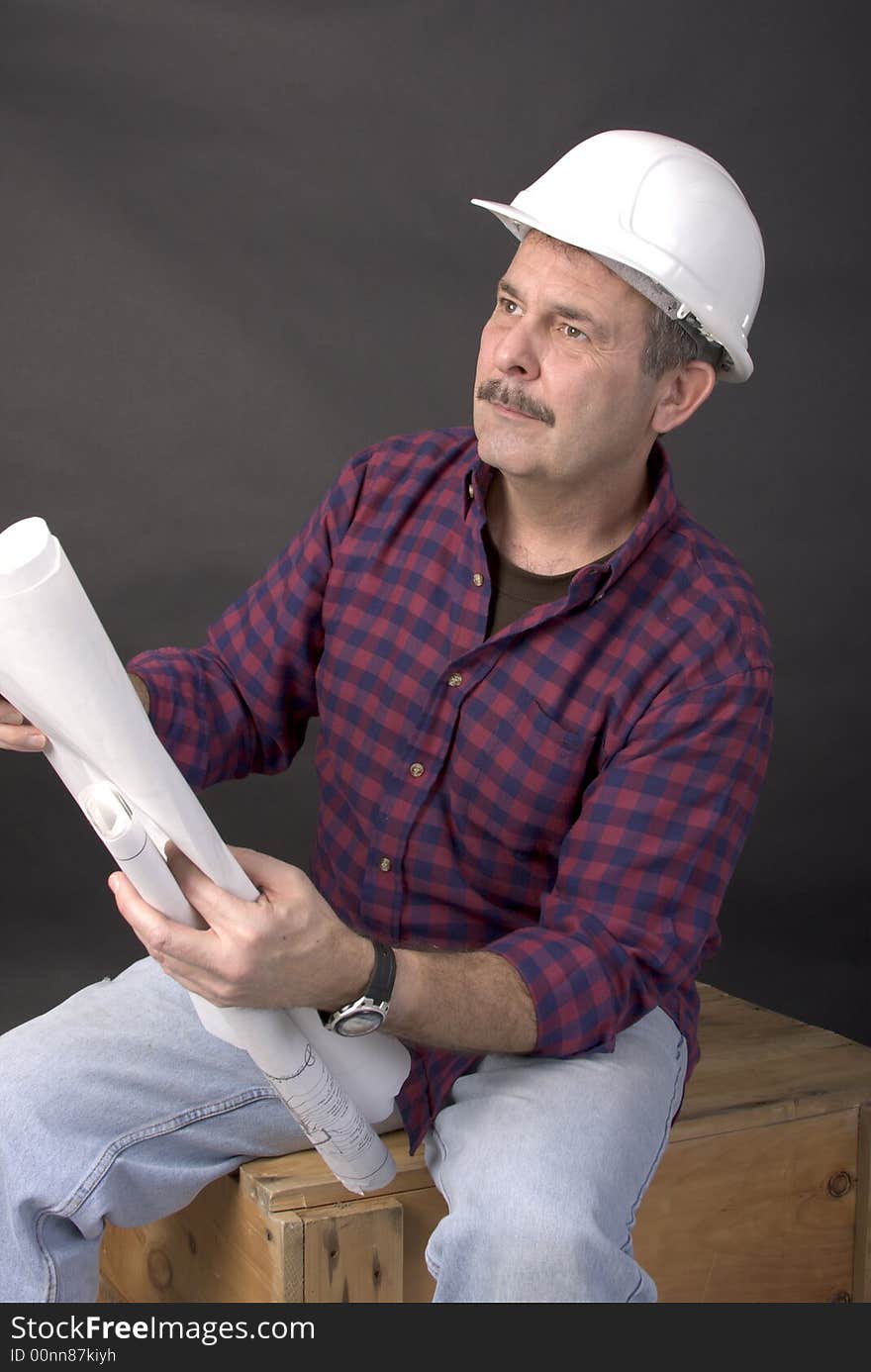 Middle-aged man with hardhat on holding blueprints in his hands while sitting on a wooden crate. Middle-aged man with hardhat on holding blueprints in his hands while sitting on a wooden crate.