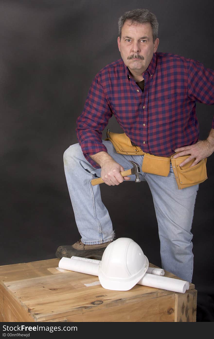 Worker holding hammer in hand with blueprints and hardhat laying on wooden crate, with black background. Worker holding hammer in hand with blueprints and hardhat laying on wooden crate, with black background