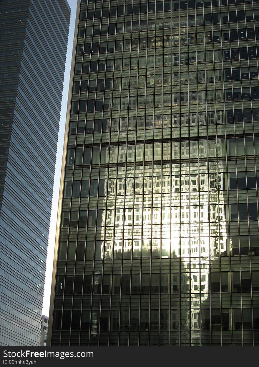 The picture display a close up of two skyscrapers. On the glassy surface of the skyscrapers on the right it is visible the reflection of another skyscraper. The picture display a close up of two skyscrapers. On the glassy surface of the skyscrapers on the right it is visible the reflection of another skyscraper