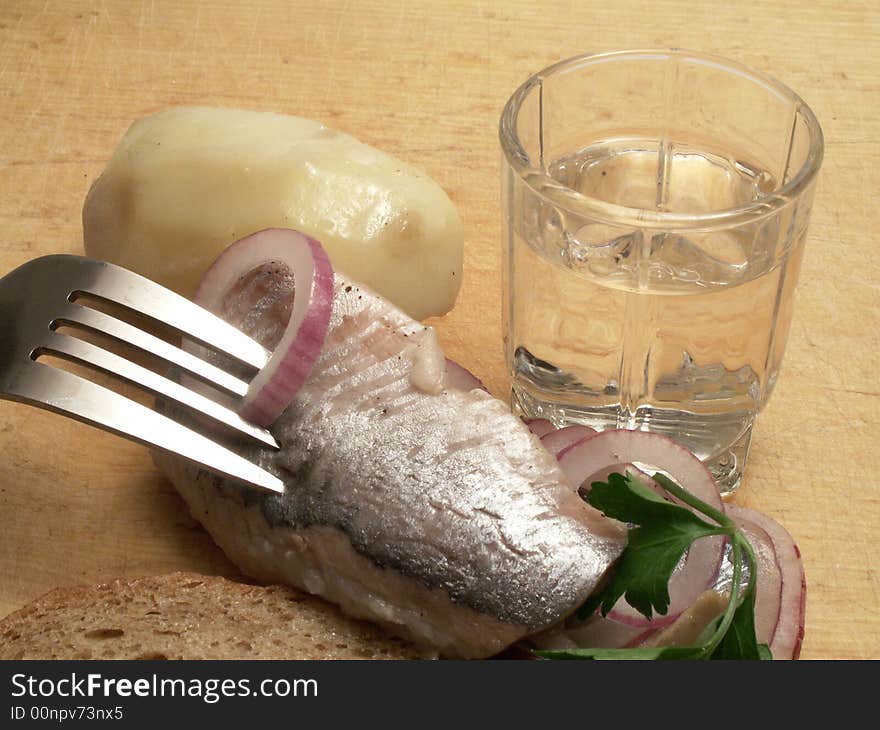 Glass of vodka, boiled potatoes and fork with herring and onion on background with wooden plate. Glass of vodka, boiled potatoes and fork with herring and onion on background with wooden plate