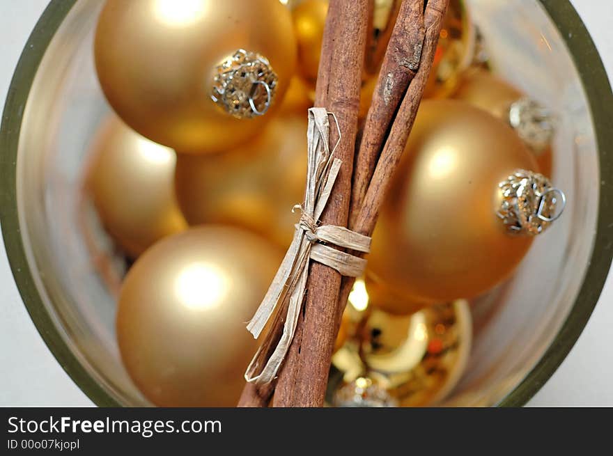 A closeup of golden Christmas balls, in a glass container with a bunch of cinnamon sticks placed across the mouth of the glass. A closeup of golden Christmas balls, in a glass container with a bunch of cinnamon sticks placed across the mouth of the glass.