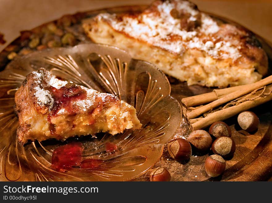 A slice of delicious homemade cake in front on the glass plate. The cake itself is in the backround together with cinnamon sticks, filbert nuts and raisins. Tha focus is on the slice of cake and nuts in the foreground.