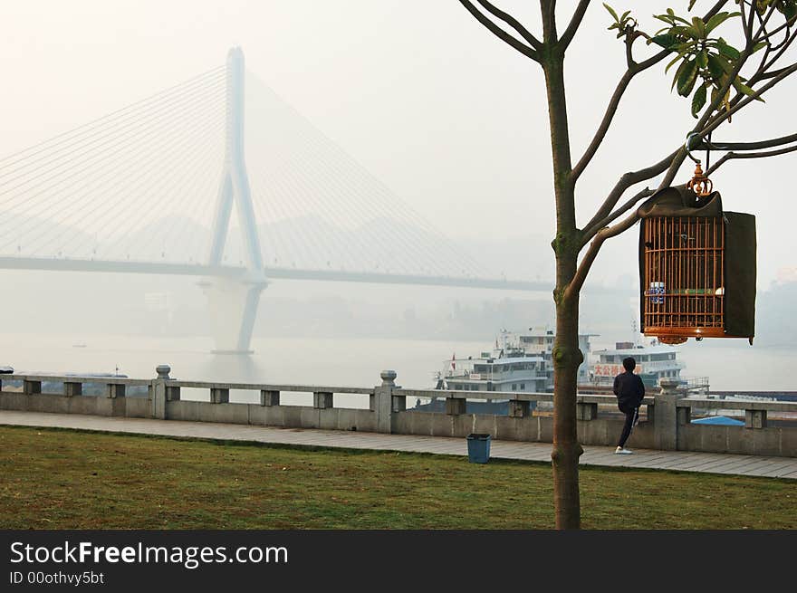 A Birdcage is hanged by Branches,nearby,a women is doing Physical Exercise in the edge of Yangtze River in the morning. A Birdcage is hanged by Branches,nearby,a women is doing Physical Exercise in the edge of Yangtze River in the morning.