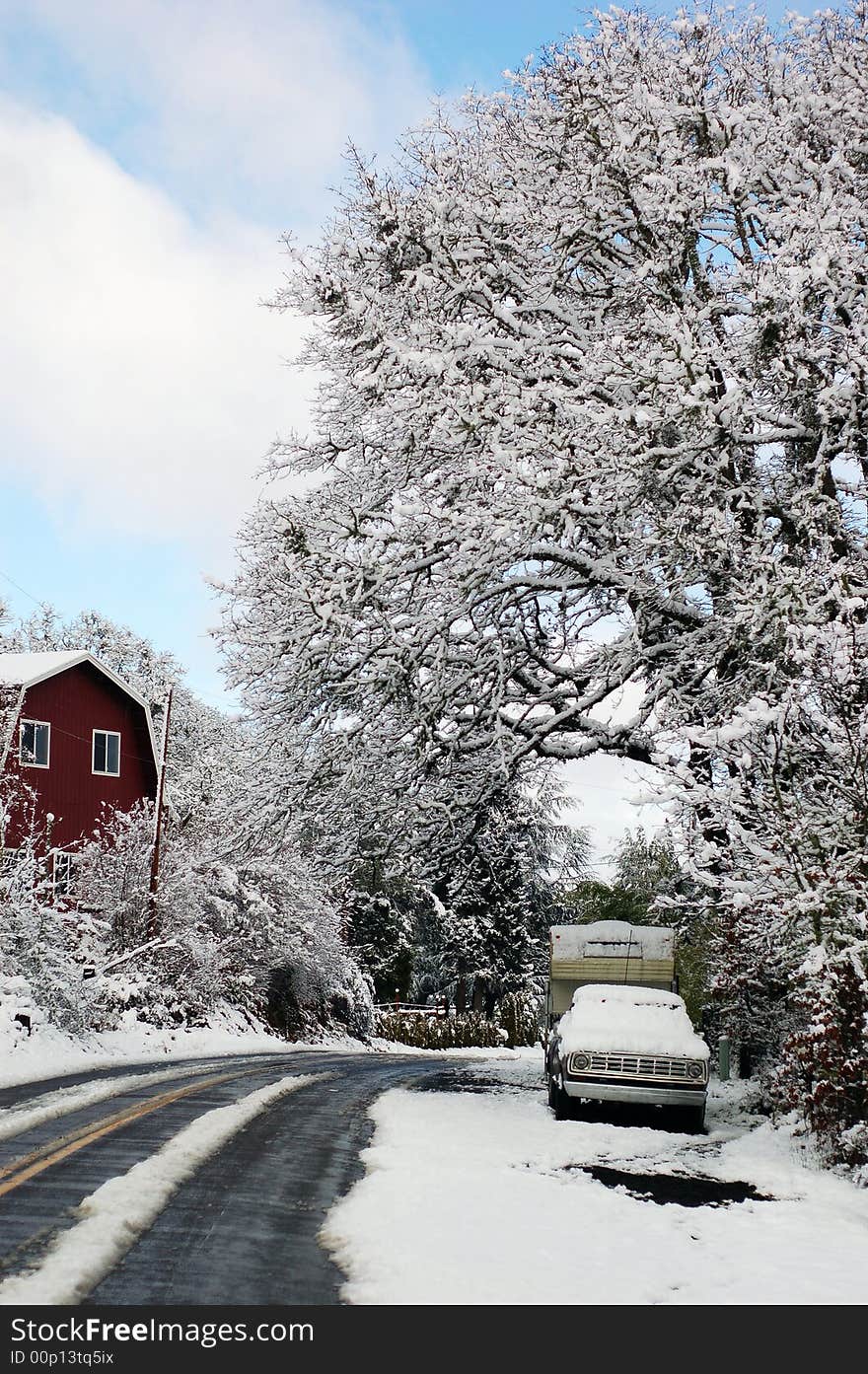 A truck under a tree on the side of a road, covered in snow at day time with a red barn to the left. A truck under a tree on the side of a road, covered in snow at day time with a red barn to the left.