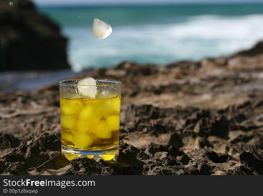 Tumbler glass with ice falling into the glass containing whiskey with the ocean and blue sky in the background. Tumbler glass with ice falling into the glass containing whiskey with the ocean and blue sky in the background