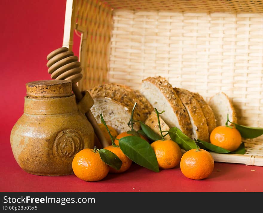 Still life of a honey pot with dipper, tangerines  with leaves , and a loaf of bread in a wicker basket, on red background illuminated by the morning sun from the side. Still life of a honey pot with dipper, tangerines  with leaves , and a loaf of bread in a wicker basket, on red background illuminated by the morning sun from the side