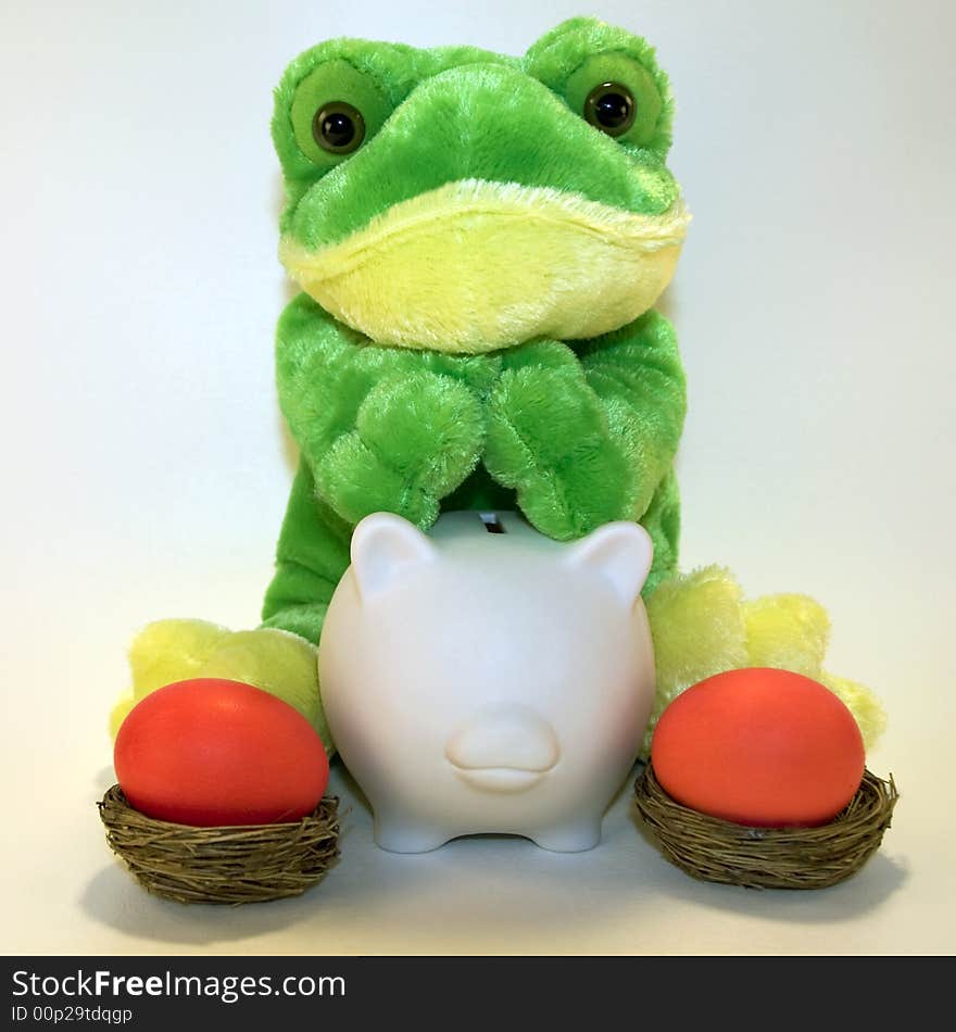 Cute plush frog sitting behind a white ceramic piggy bank with two red Easter eggs in nests. Cute plush frog sitting behind a white ceramic piggy bank with two red Easter eggs in nests.