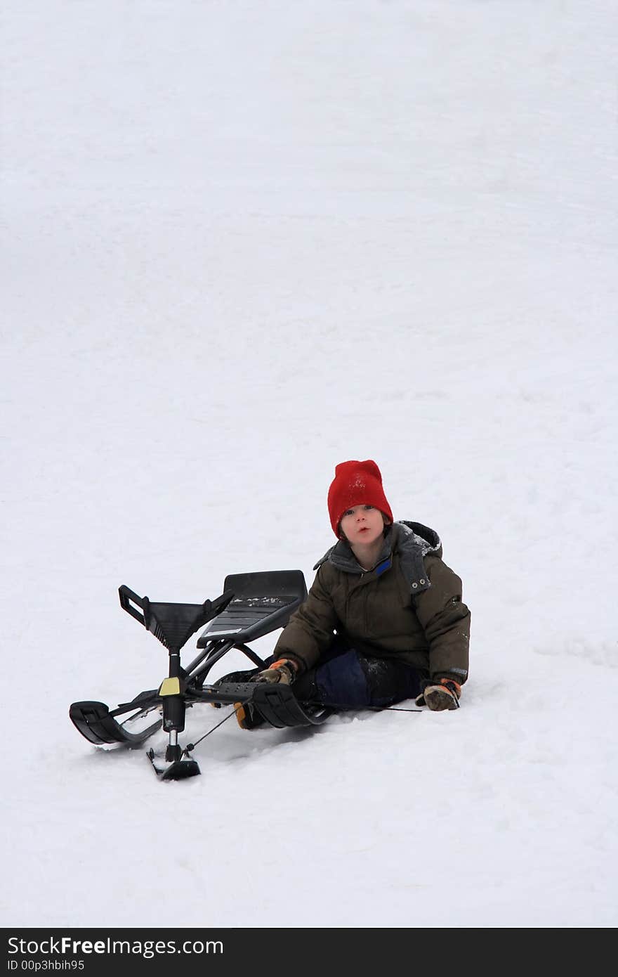 A young boy enjoying a winter day resting at the bottom of a steep toboggan hill with his sled