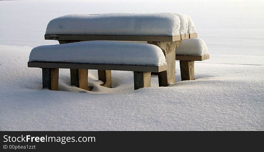 A lakeside picnic table and benches are covered with snow. A lakeside picnic table and benches are covered with snow.