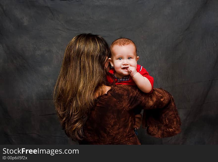 Mother dressed in brown from the back is holding her baby boy with sparkling eyes who's sucking his thumb, having a mischievous look and facing camera, isolated on a dark background. Mother dressed in brown from the back is holding her baby boy with sparkling eyes who's sucking his thumb, having a mischievous look and facing camera, isolated on a dark background