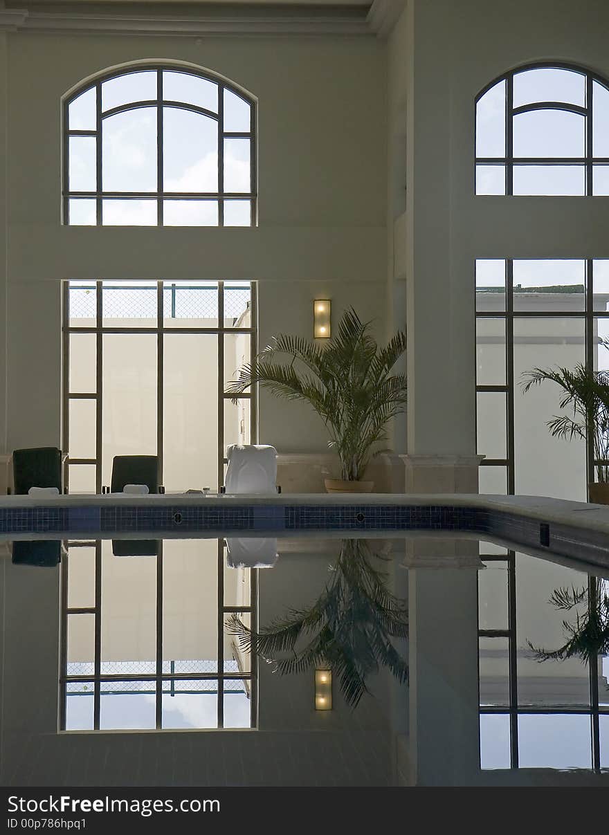 Lounge chairs and potted palm tree reflected with windows in a quiet indoor pool in a spa. Lounge chairs and potted palm tree reflected with windows in a quiet indoor pool in a spa