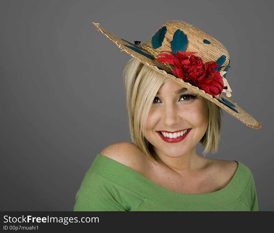 A blonde model with brilliant smile in a flowered straw hat on grey background. A blonde model with brilliant smile in a flowered straw hat on grey background