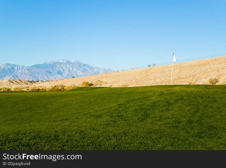 An image of a golf course in a desert with a flag in a foreground and mountains in the background.