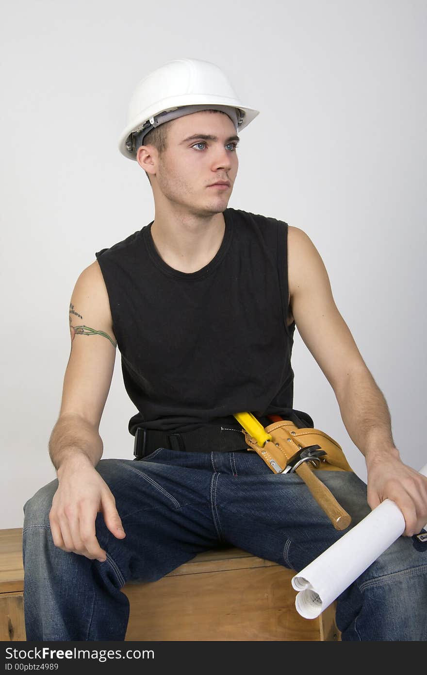 Young man wearing hardhat and toolbelt, sitting on a wooden crate and holding a set of blueprints. Young man wearing hardhat and toolbelt, sitting on a wooden crate and holding a set of blueprints.