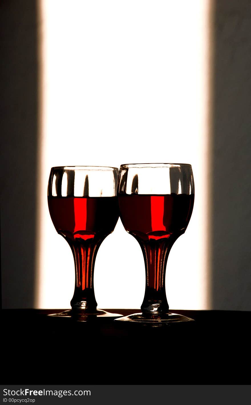 Wine glasses stand with a bottle of wine dramatically backlit to show of the beauty of the color of the containers’ contents. Wine glasses stand with a bottle of wine dramatically backlit to show of the beauty of the color of the containers’ contents.