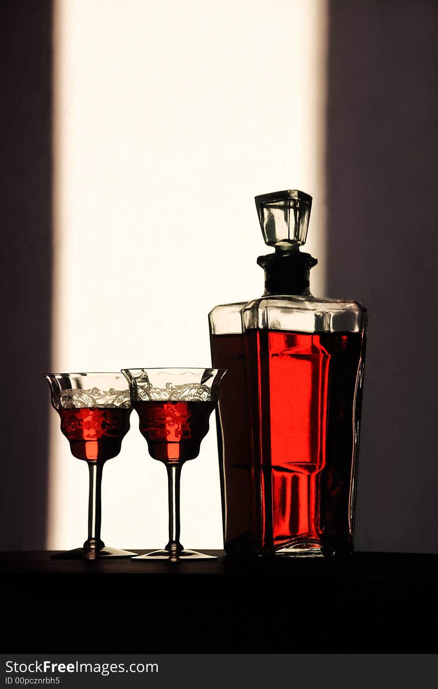 Wine glasses stand with a bottle of wine dramatically backlit to show of the beauty of the color of the containers’ contents. Wine glasses stand with a bottle of wine dramatically backlit to show of the beauty of the color of the containers’ contents.