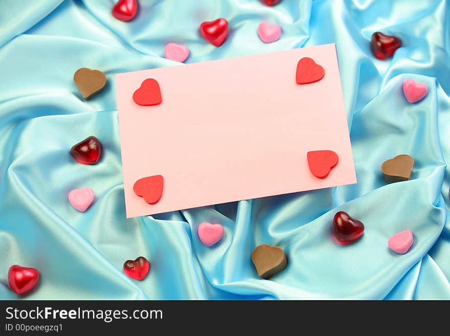 A pink valentine card lying on blue satin fabric with little hearts of different materials.