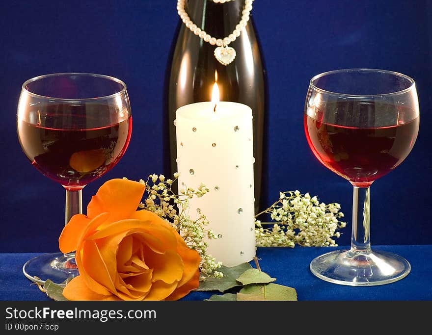 A closeup view of two glasses filled with red wine, a white candle and a peach rose with a bottle of wine and baby's breath in the background. A closeup view of two glasses filled with red wine, a white candle and a peach rose with a bottle of wine and baby's breath in the background.