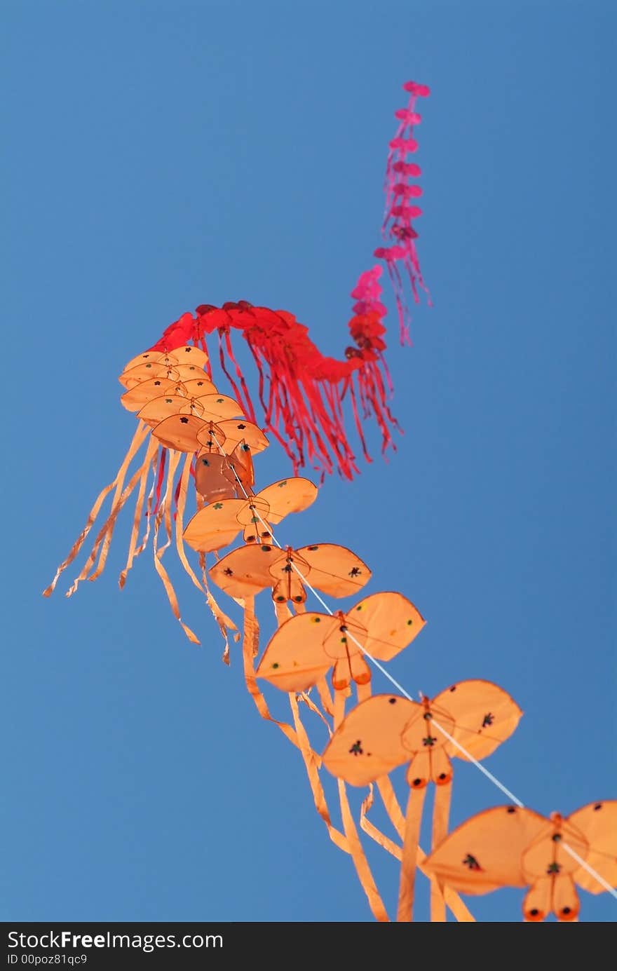 String of kites in the colours orange, red and pink on a blue sky background. Shallow depth of field with the nearest kites in focus.