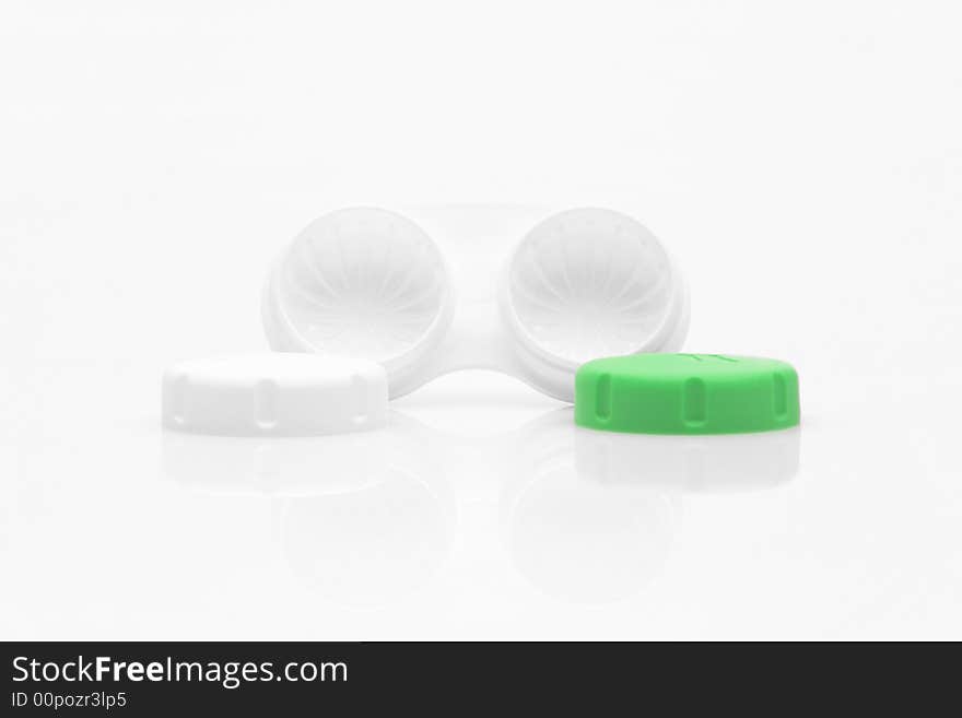 A white contact lens case on a white surface. One of the compartment lids is lime green. The lids are placed in front of the holder. A white contact lens case on a white surface. One of the compartment lids is lime green. The lids are placed in front of the holder