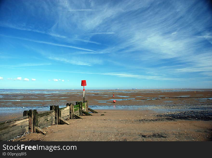 Estuary beach at low tide showing very large area of sand, groyne or breakwater, and red danger marker set against dramatic sky showing high clouds and con trails