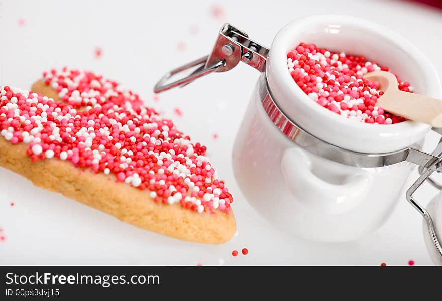 Red, white and pink nonpareils on heart-shaped cookie and in white porcelin storage jar. Red, white and pink nonpareils on heart-shaped cookie and in white porcelin storage jar.