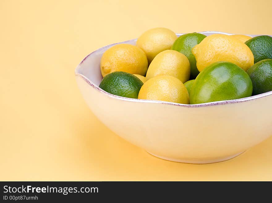 A bowl of lemons and limes in a white bowl on a yellow background. A bowl of lemons and limes in a white bowl on a yellow background.