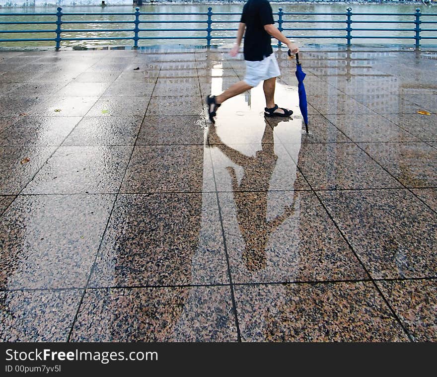 A young man strolling along the wet walkway by a river with an umbrella after a late afternoon shower. A young man strolling along the wet walkway by a river with an umbrella after a late afternoon shower