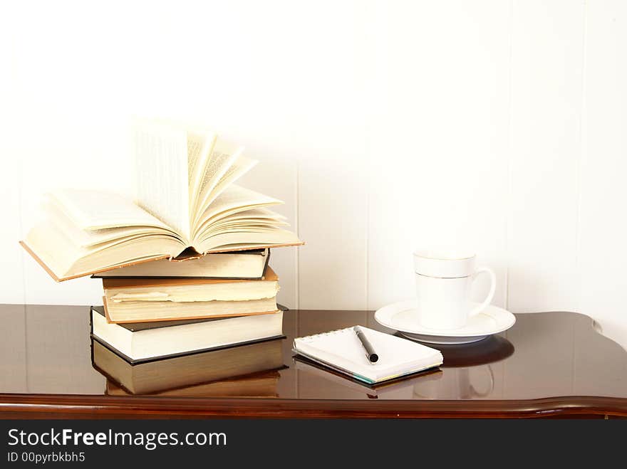 Working or studying. Books stacked on polished dark cherry wood table with notepad, pen and cofee.