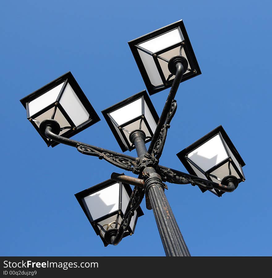 Three ornate street lamps on a lamp post against a blue sky in Romania