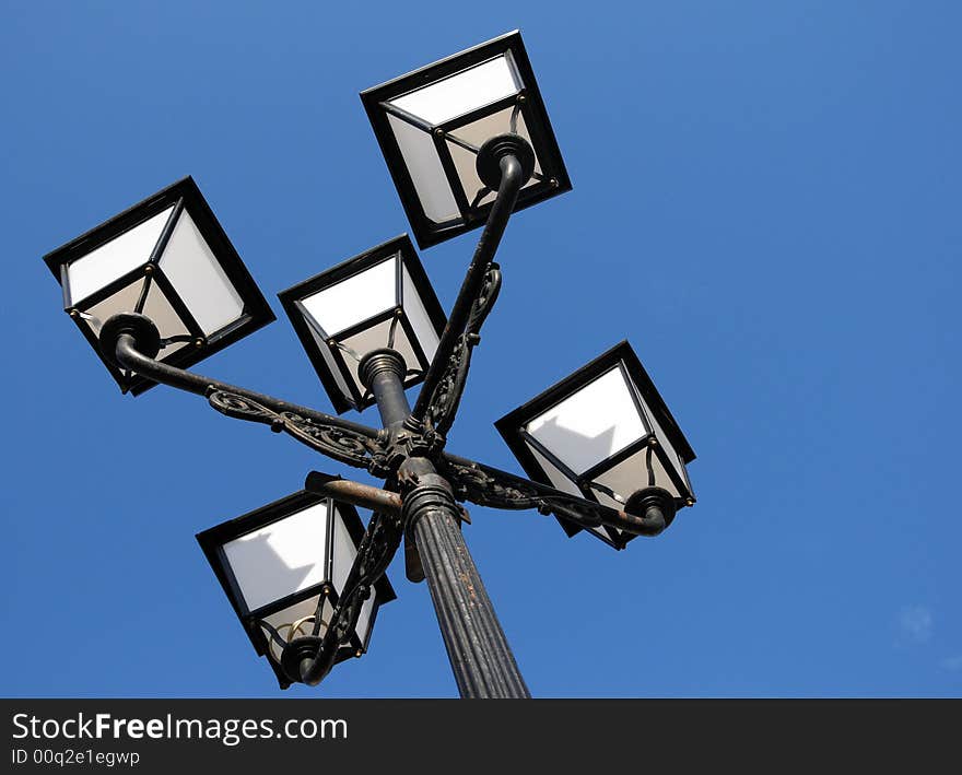 Three ornate street lamps on a lamp post against a blue sky in Romania