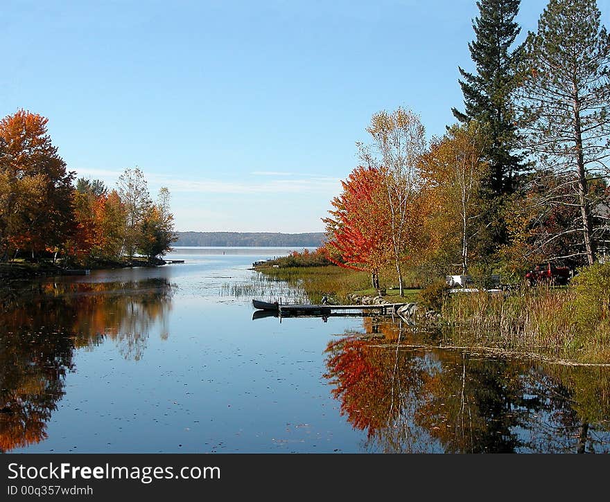 Brilliant tree-lined shore reflected in halcyon waters of small inlet. Small pier and boat along shoreline. Brilliant tree-lined shore reflected in halcyon waters of small inlet. Small pier and boat along shoreline.