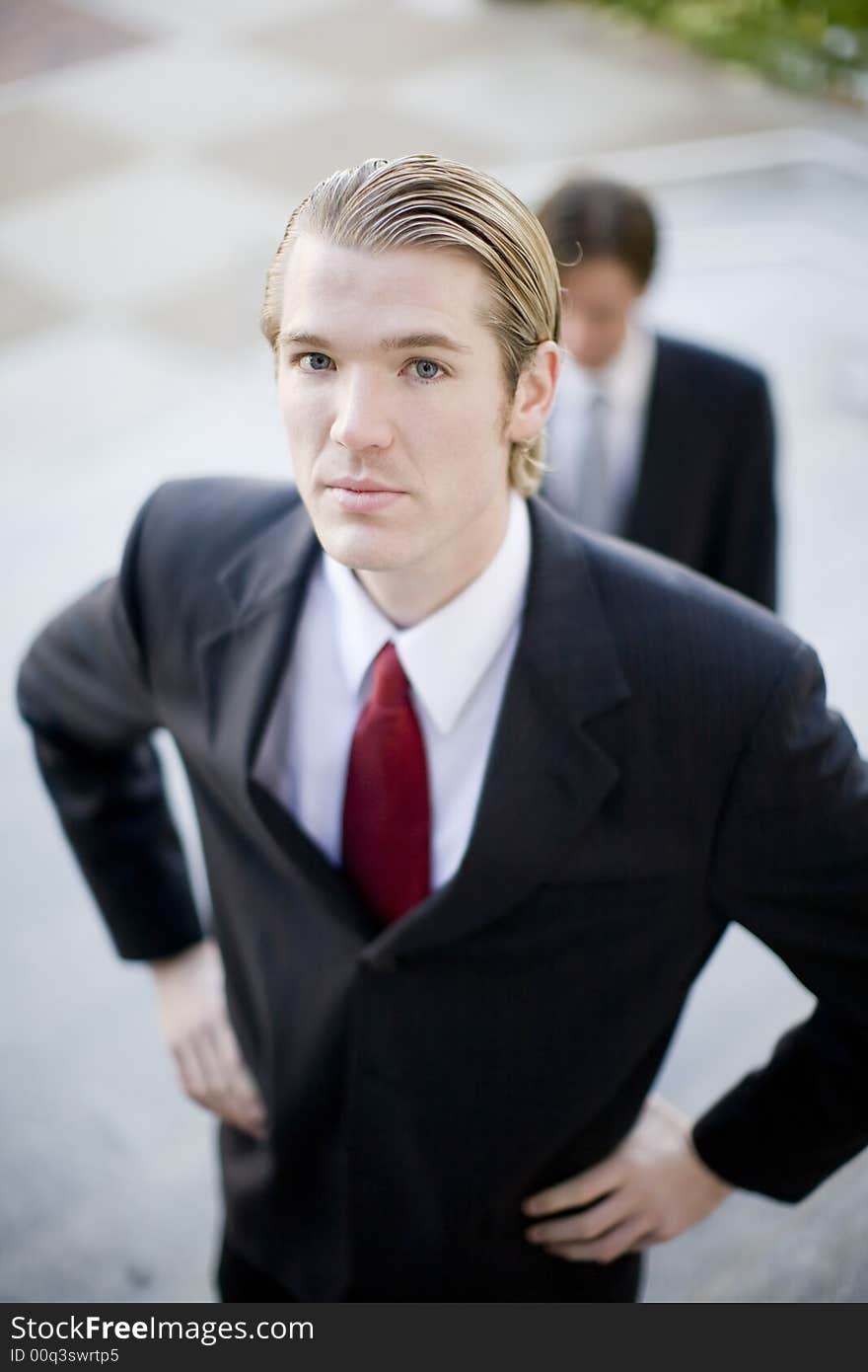 Two businessmen standing on steps one with arms on hips the other behind the first looking down