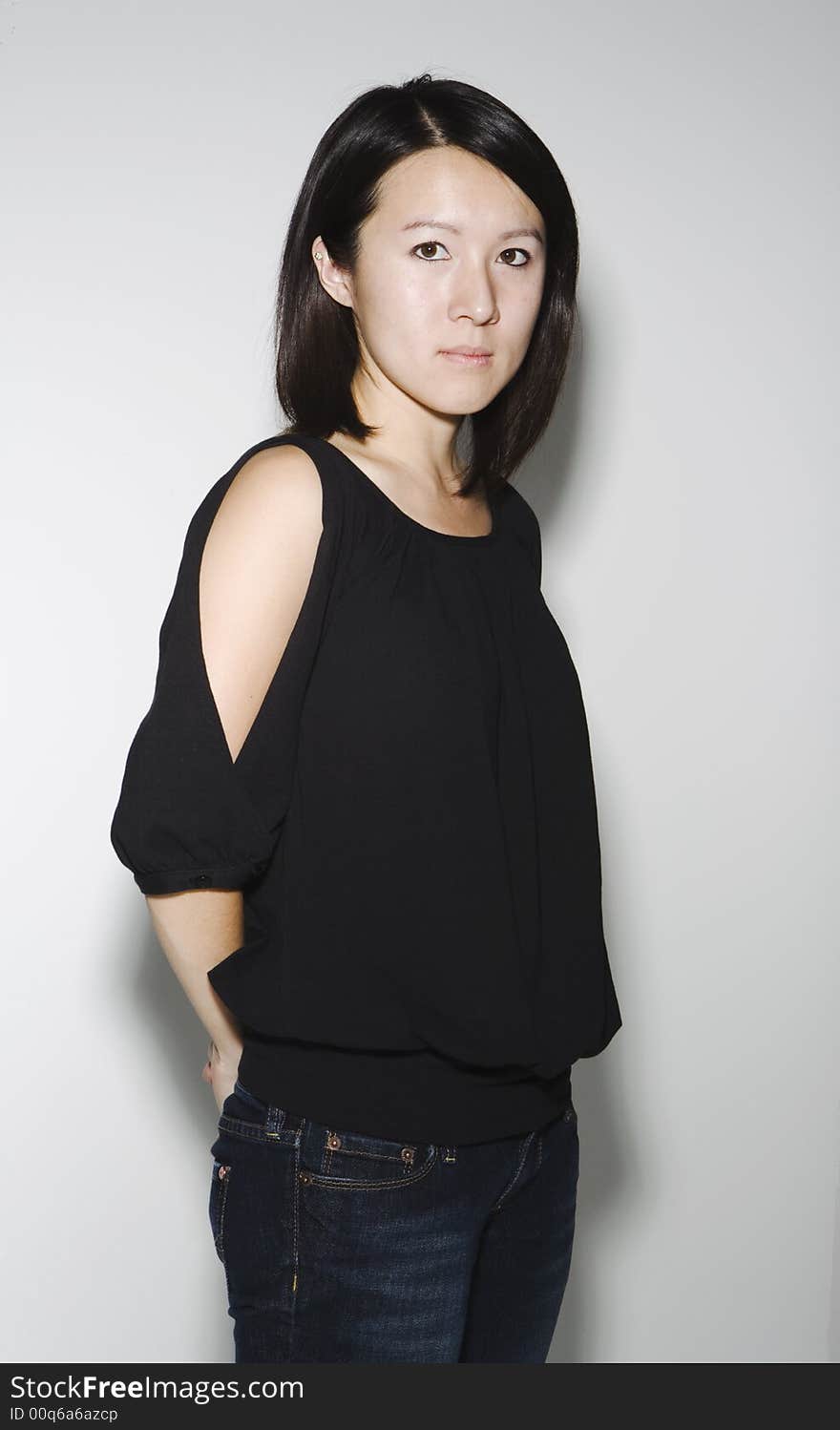 Asian woman against a white wall look directly into camera with her hand behind her back.  She is wearing a black top and denim jeans. Asian woman against a white wall look directly into camera with her hand behind her back.  She is wearing a black top and denim jeans.