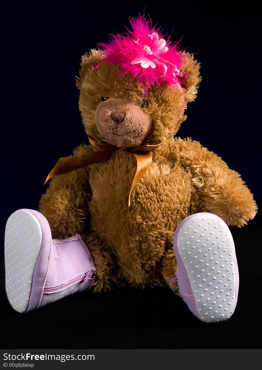 A brown teddybear with pink boots and pink feathers on its head. Black background.