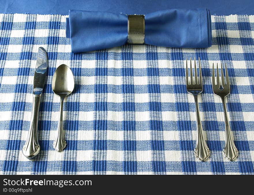 Silverware and blue napkin with silver napkin ring lay on a checkered blue dinner placemat. Silverware and blue napkin with silver napkin ring lay on a checkered blue dinner placemat.