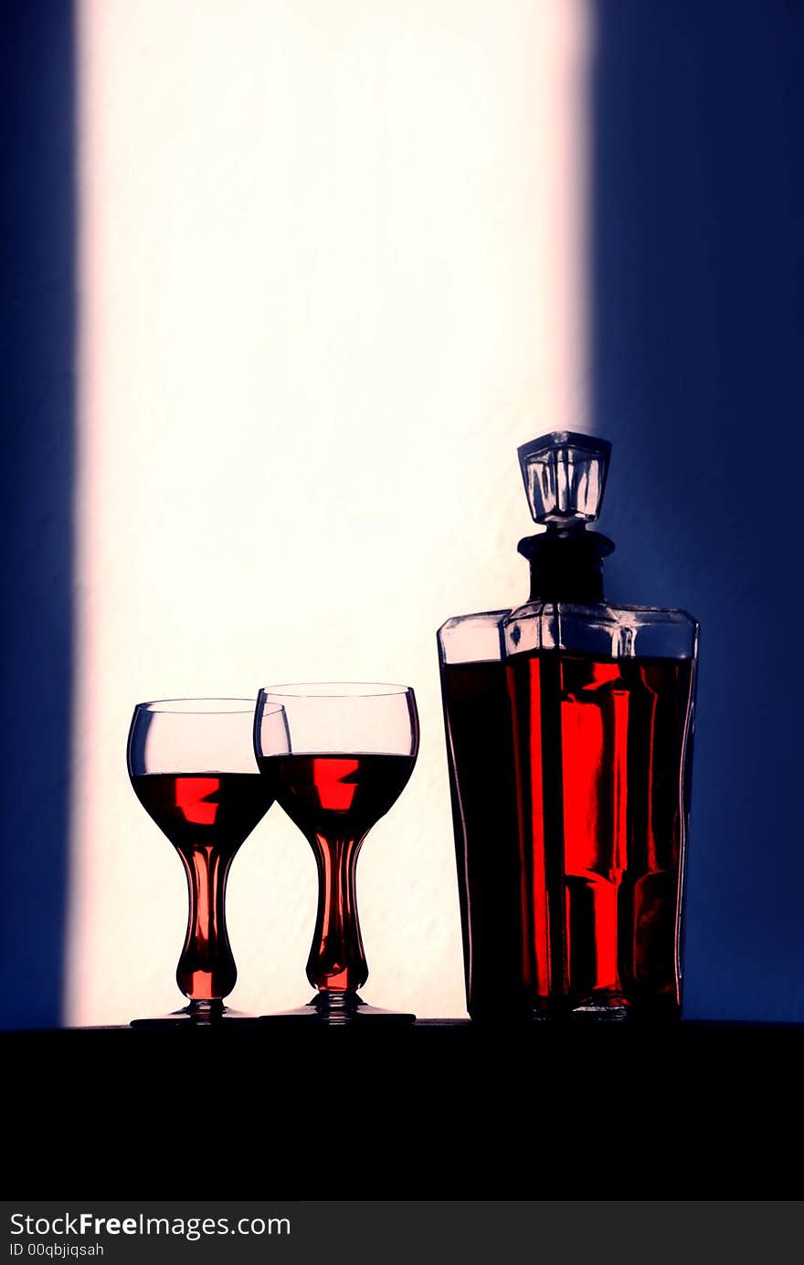 Wine glasses stand with a bottle of wine dramatically back lit to show of the beauty of the color of the containers’ contents.