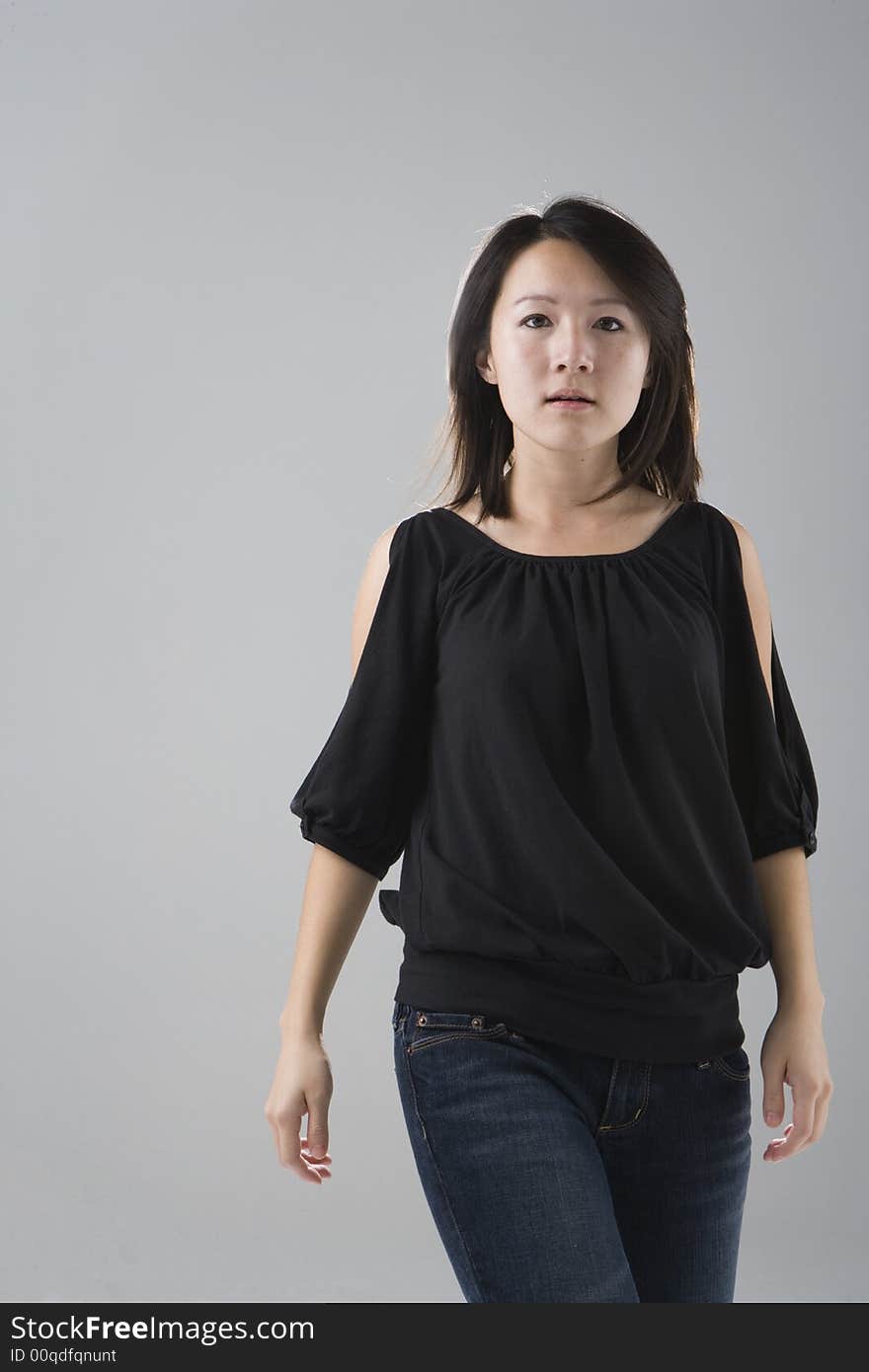 Young Asian woman wearing a black top and denim jeans walks forward with her eyes looking at the viewer against a gray studio background. Young Asian woman wearing a black top and denim jeans walks forward with her eyes looking at the viewer against a gray studio background.