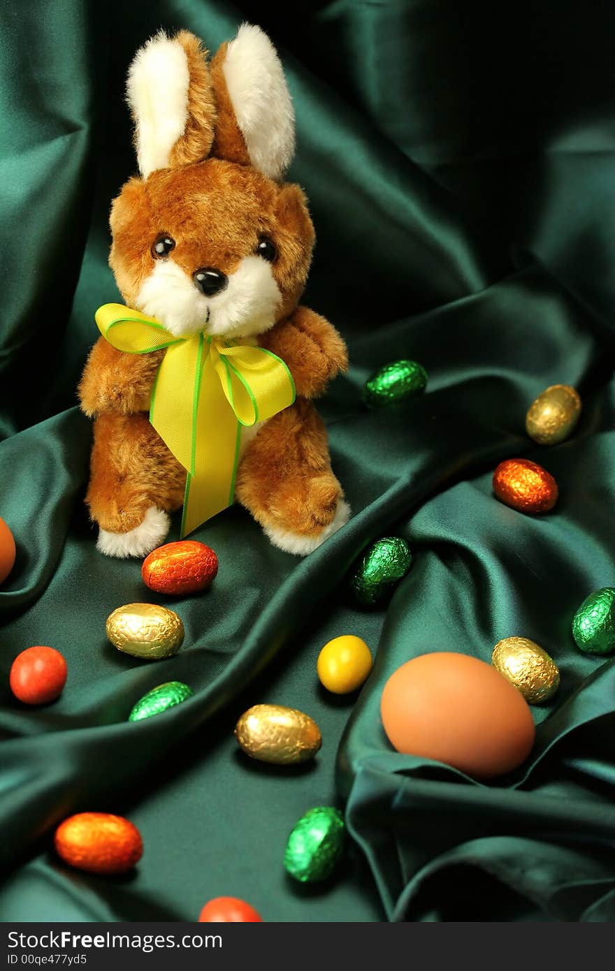 Easter theme - a fluffy bunny (rabbit) with a yellow ribbon sitting on a dark green satin fabric - textile, surrounded by colorful Easter eggs. Easter theme - a fluffy bunny (rabbit) with a yellow ribbon sitting on a dark green satin fabric - textile, surrounded by colorful Easter eggs.